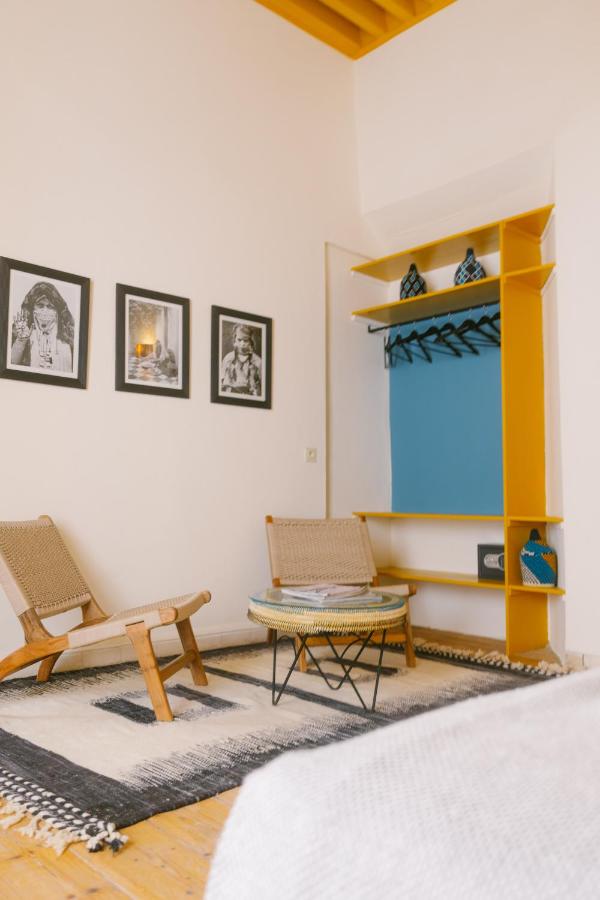 10 BEST Riads in Essaouira 10 A cozy room with two wooden chairs and a round table on a patterned rug evokes the charm of riads in Essaouira. Three black-and-white photos decorate the wall, while a yellow open closet with blue accents holds vases and hangers.
