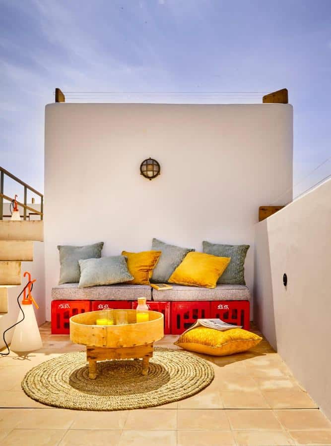 10 BEST Riads in Essaouira 23 A cozy outdoor patio, reminiscent of riads in Essaouira, features a cushioned bench with colorful pillows, a round woven rug, a small wooden table with drinks, and a floor lamp on tan tiles against a white wall under a clear blue sky.
