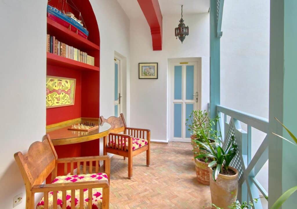 10 BEST Riads in Essaouira 33 A cozy balcony inspired by riads in Essaouira, featuring wooden chairs and a round table, a red arch bookshelf with books and a model ship, potted plants, colorful cushions, a hanging lantern, and doors with a framed picture on the wall.