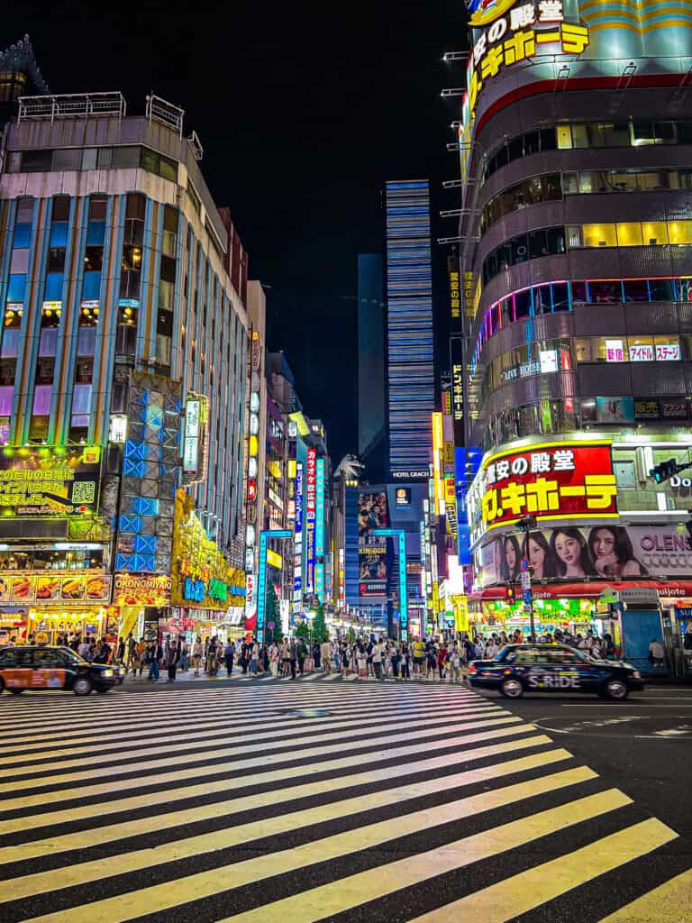 ULTIMATE 10 Day Japan Travel Itinerary 5 A busy city street at night in Tokyo, Japan, with colorful neon signs, tall buildings, a large crosswalk, cars passing by, and many people—just one of the vibrant sights you’ll experience on a 10 day Japan travel itinerary.