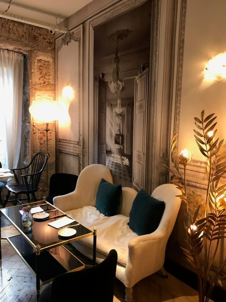 15 Most Fun Restaurants in Madrid 45 A cozy sitting area with a white loveseat, dark green pillows, a glass coffee table, gold decorative plant, and a mural of an ornate hallway evokes the charm of fun restaurants in Madrid, all illuminated by warm lamps.