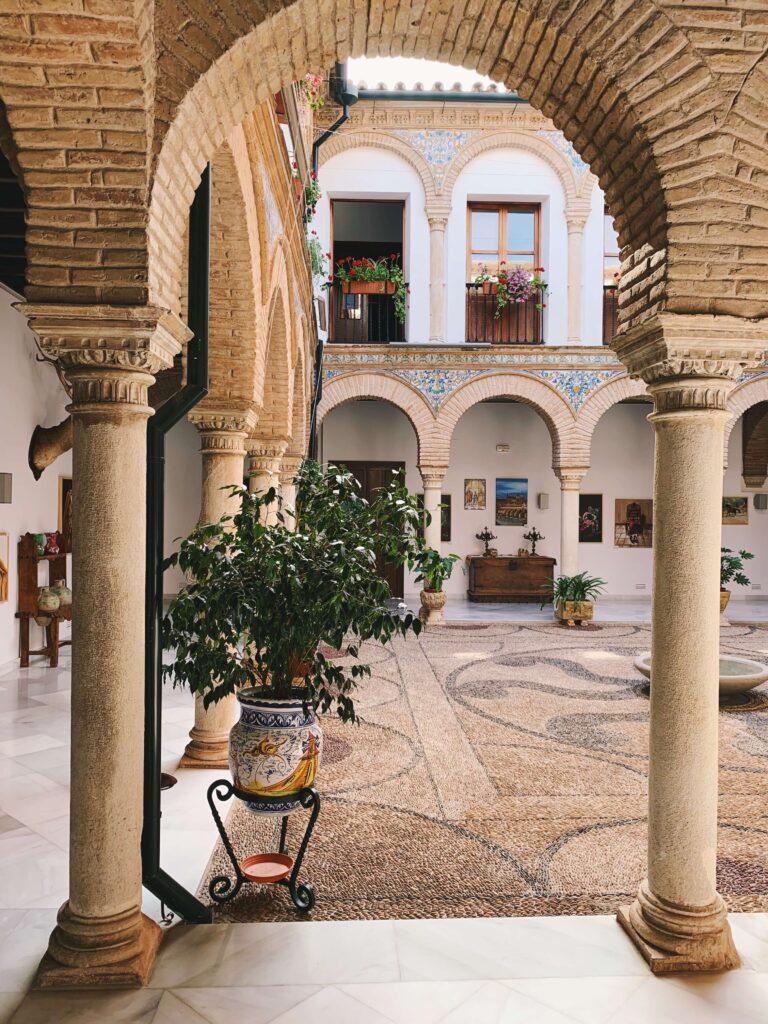 Southern Spain Itinerary 10 Days 11 A sunlit Andalusian courtyard, perfect for your Southern Spain itinerary 10 days, with stone arches, columns, patterned tile floors, a central potted plant, wall art, and a balcony adorned with flower pots and intricate tiles.