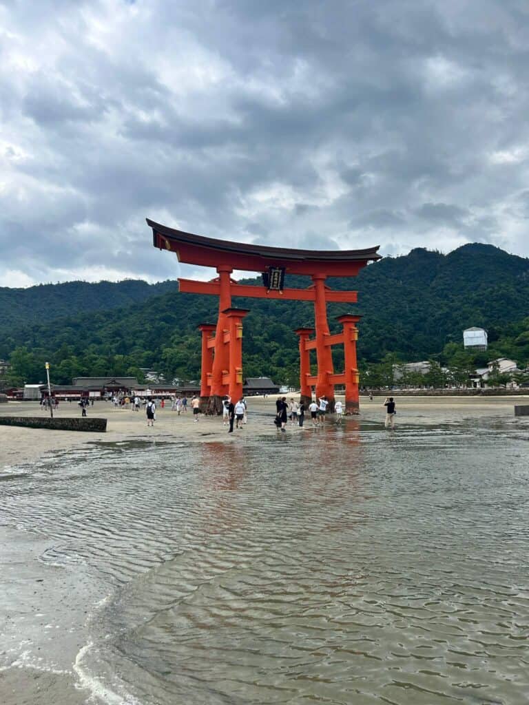 ULTIMATE 10 Day Japan Travel Itinerary 19 Tourists walk near the large, iconic red torii gate of Itsukushima Shrine—an unmissable stop on any 10 day Japan travel itinerary—with green forested hills and a cloudy sky in the background.