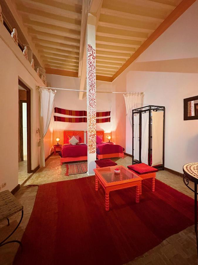 10 BEST Riads in Essaouira 46 A cozy bedroom inspired by riads in Essaouira, with warm lighting, a double bed dressed in red bedding, matching rugs, a low orange table, two chairs, a folding screen, and decorative textiles on the walls and above the bed.
