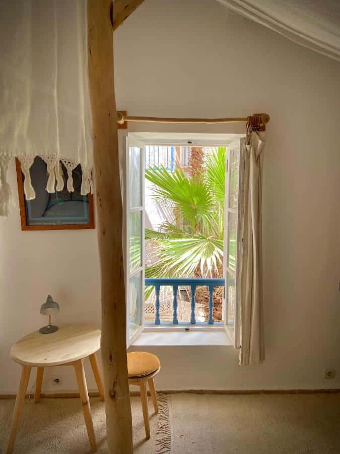 10 BEST Riads in Essaouira 39 A bright, cozy room inspired by riads in Essaouira features a small round wooden table, a matching stool, and a lamp by an open window. Green palm leaves and neutral tones create a calming atmosphere filled with natural sunlight.