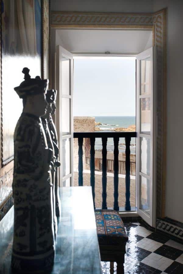 10 BEST Riads in Essaouira 27 A sunlit room in one of the riads in Essaouira, with ornate blue-and-white ceramic statues on a table, an open door leading to a balcony, and views of the sea and rocky shore in the distance.