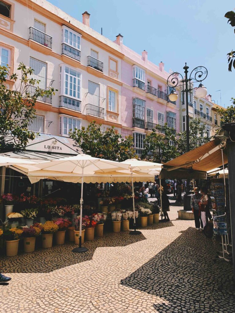 Southern Spain Itinerary 10 Days 14 A flower stall with colorful bouquets under white umbrellas sits on a cobblestone street, surrounded by trees and pastel-colored buildings—an inviting stop on a Southern Spain itinerary 10 days—with people strolling and enjoying the lively scene.