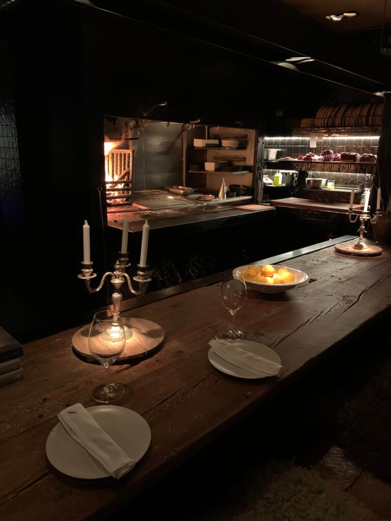 15 Most Fun Restaurants in Madrid 46 A rustic wooden table is set with plates, glasses, napkins, candles, and a bowl of lemons. In the background of one of the fun restaurants in Madrid, an open kitchen with a wood-fired oven glows warmly amid shelves of ingredients and cookware.
