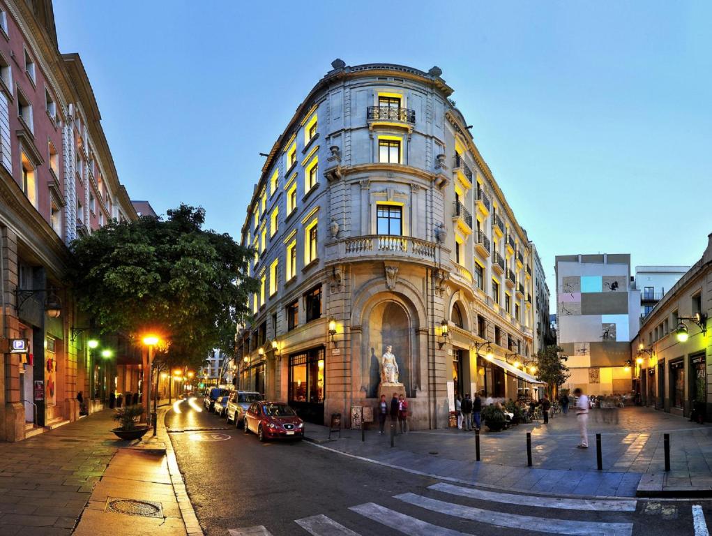 Top Barcelona Hotels with Rooftop Pool 35 A grand, historic building with illuminated windows sits at a street corner at dusk, reminiscent of Barcelona hotels with rooftop pool views. A statue graces its entrance while the lively street bustles with trees, parked cars, and pedestrians.