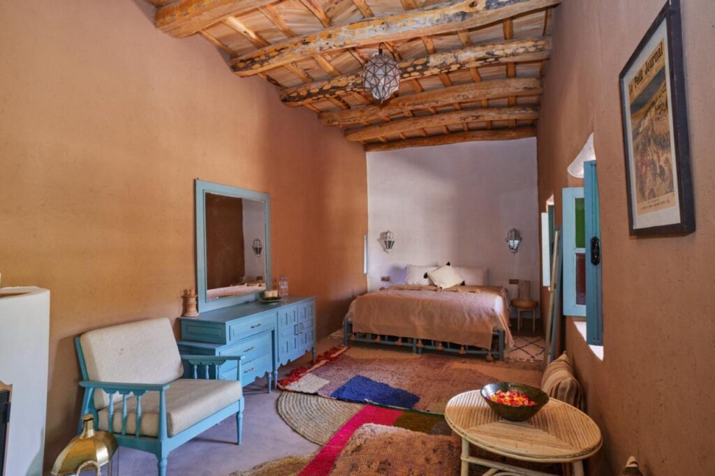 8 SPECTACULAR Agafay Desert Camps 24 Cozy rustic bedroom with a wooden ceiling, beige walls, a large bed, blue furniture, colorful woven rugs, and wicker chairs with a small table evokes the inviting charm of Agafay Desert camps as natural light streams through the window.