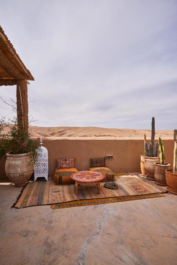 8 SPECTACULAR Agafay Desert Camps 25 A cozy desert patio with colorful cushions, a round patterned table, large potted plants, and cacti on a rug evokes the charm of Agafay Desert camps, overlooking sandy dunes under a partly cloudy sky.