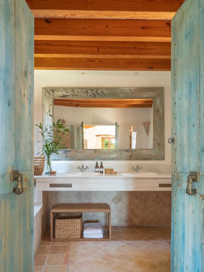 Unique Agroturismos in Mallorca 13 A rustic bathroom inspired by agroturismos in Mallorca, with double sinks, a large distressed wooden mirror, blue weathered doors, wicker baskets, and a potted plant, featuring tiled floors and exposed wooden ceiling beams.