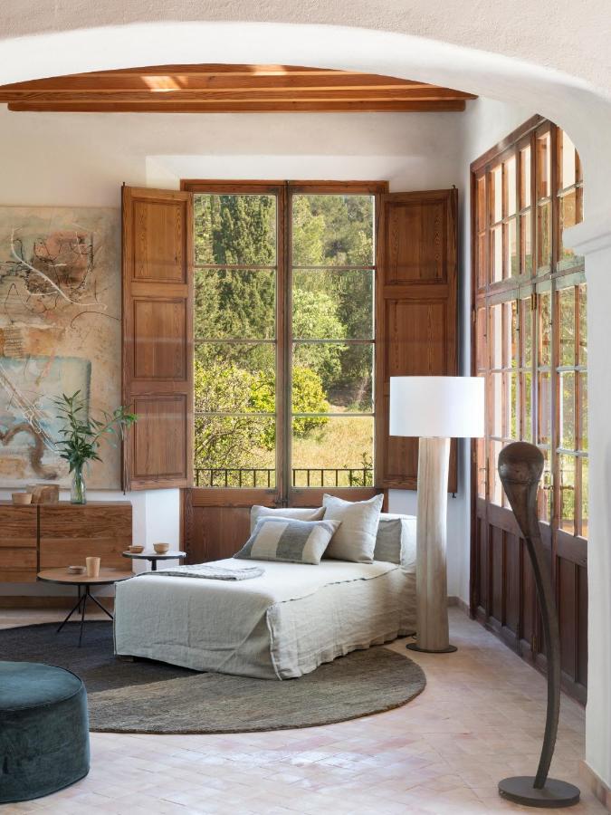 Unique Agroturismos in Mallorca 11 A bright, cozy living room inspired by agroturismos in Mallorca, with large wooden windows, a chaise lounge with light cushions, a round rug, wooden cabinets, abstract artwork, indoor plants, and two modern floor lamps. Trees are visible outside.