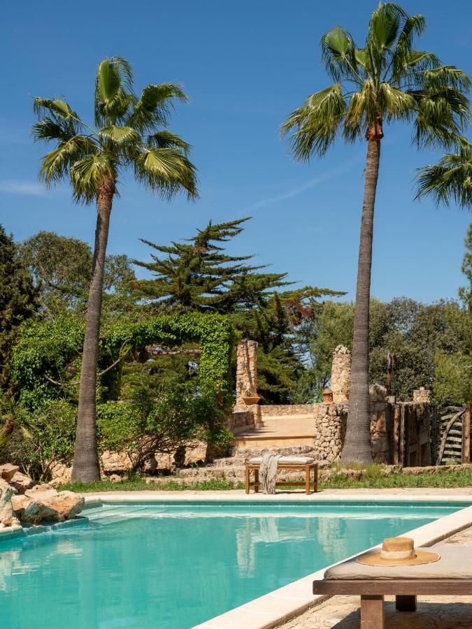 Unique Agroturismos in Mallorca 10 A serene outdoor swimming pool at one of the agroturismos in Mallorca, with clear blue water, tall palm trees, a wooden bench, and a straw hat on a towel. Lush greenery and rustic stone structures complete this tranquil escape.