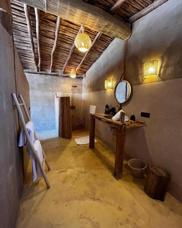 8 SPECTACULAR Agafay Desert Camps 29 A rustic bathroom inspired by agafay desert camps features a wooden ceiling, beige plaster walls, a wooden sink counter with a round mirror, wicker light fixtures, a towel ladder, and an open shower area with a log partition.