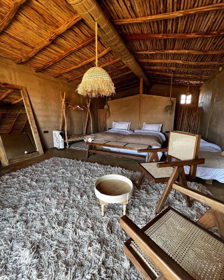 8 SPECTACULAR Agafay Desert Camps 27 A cozy rustic bedroom inspired by agafay desert camps, with wooden furniture, a large bed with white bedding, a shaggy rug, woven chairs, a round wooden table, and earthy walls and ceiling made of natural wood.
