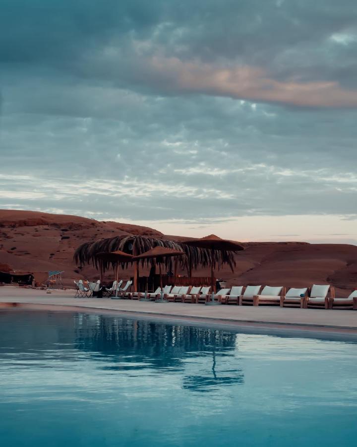 8 SPECTACULAR Agafay Desert Camps 30 A tranquil pool reflects cloudy skies, with lounge chairs along its edge. In the background, a rustic palm-thatched shelter on sandy terrain and desert dunes evokes the serene oasis charm of Agafay Desert camps.