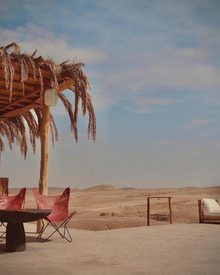 8 SPECTACULAR Agafay Desert Camps 28 A rustic patio with two red butterfly chairs, a round black table, and a wooden pergola overlooks the vast sands near Agafay desert camps under a partly cloudy sky.