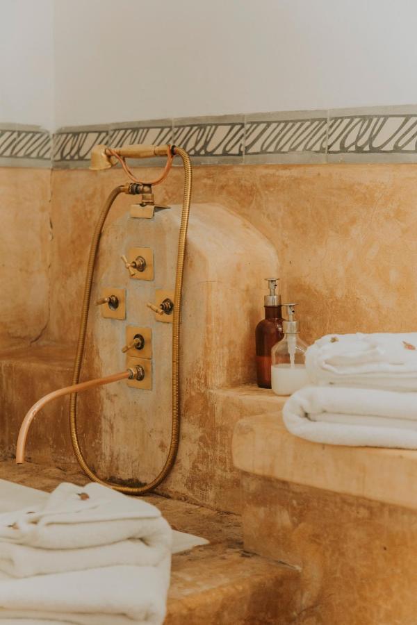 12 Charming Budget Riads in Marrakech 47 A traditional stone bath area in the style of budget riads in Marrakech, with folded white towels, two soap dispensers, and an ornate brass shower hose mounted on the wall, featuring decorative tile trim above.
