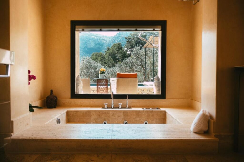 Unique Agroturismos in Mallorca 20 A sunken bathtub in a warm-toned bathroom faces a large window with a scenic view of trees, mountains, and an outdoor seating area—evoking the tranquil charm found at agroturismos in Mallorca.