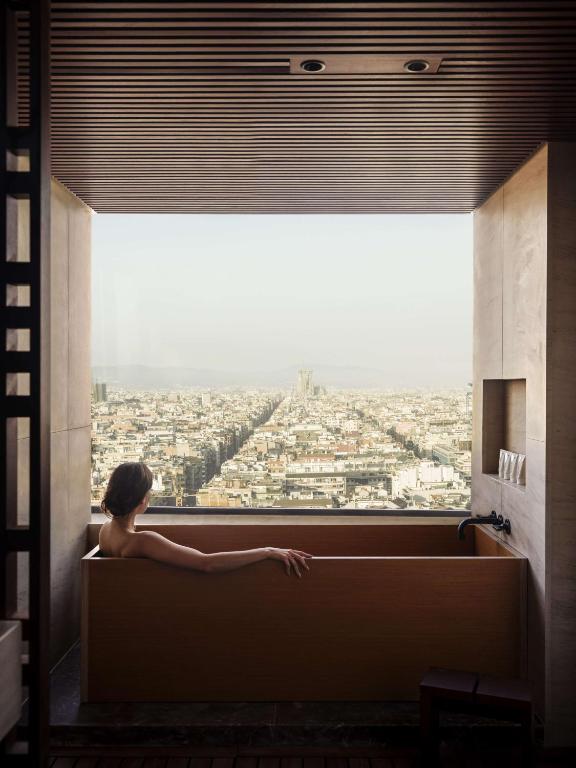Top Barcelona Hotels with Rooftop Pool 39 A person relaxes in a modern wooden bathtub by a large window, overlooking a sprawling cityscape under a clear sky—evoking the serene luxury found in top Barcelona hotels with rooftop pool views extending far into the distance.
