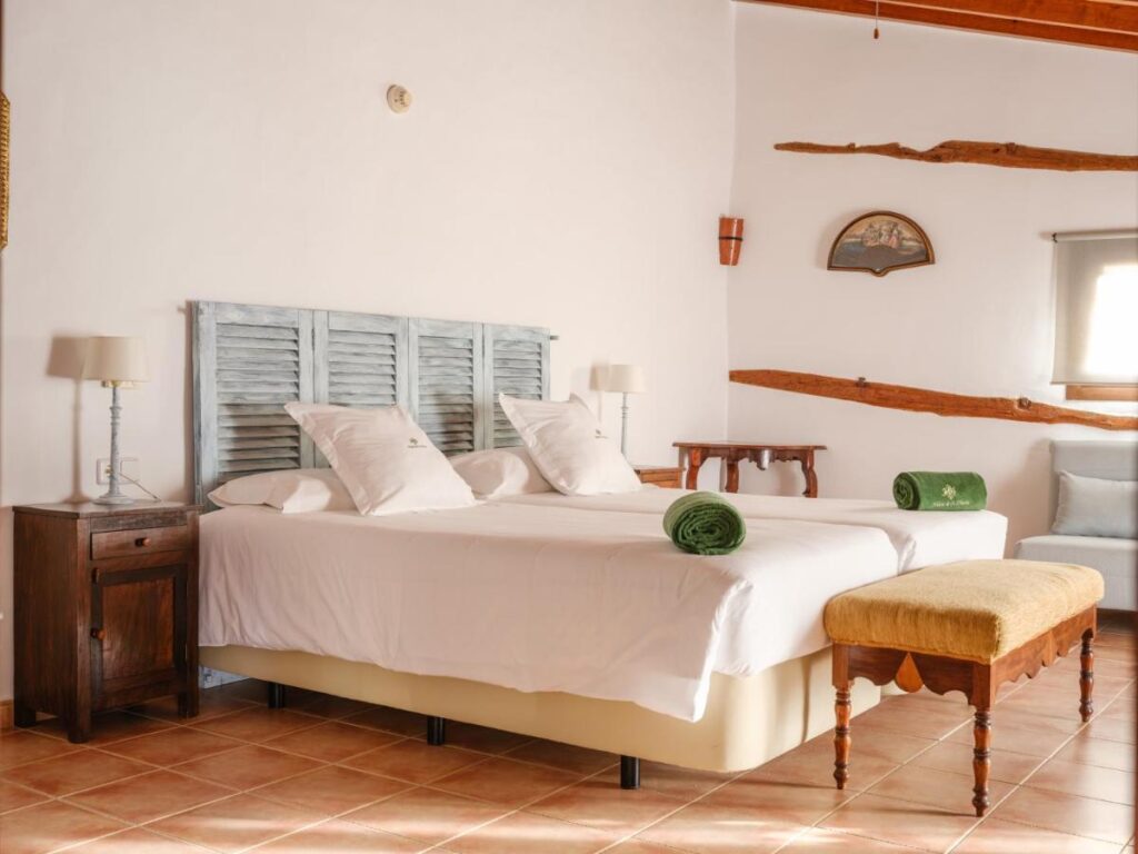 Unique Agroturismos in Mallorca 38 A cozy bedroom, typical of agroturismos in Mallorca, features two single beds pushed together, white linens, green towels, wooden furniture, a cushioned bench, and rustic wooden beams on white walls for a warm and inviting atmosphere.
