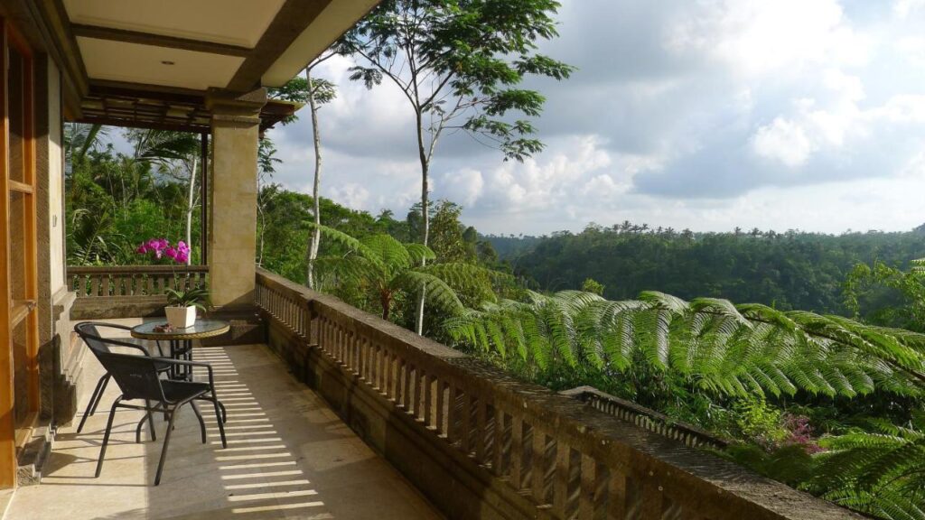 13 Most UNIQUE Places to Stay in Ubud 52 A spacious balcony with a table and two chairs overlooks lush green tropical trees and a distant forest under a partly cloudy sky—one of the unique places to stay in Ubud. Bright pink flowers add color near a wooden door to the left.
