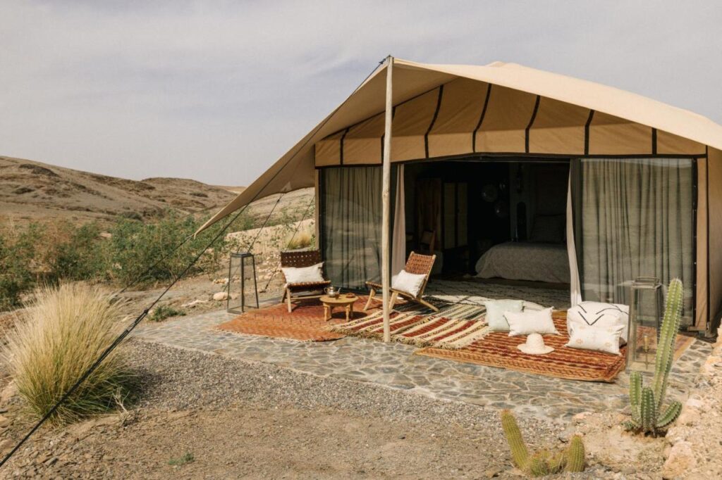 8 SPECTACULAR Agafay Desert Camps 22 A glamping tent with open canvas sides reveals a cozy bed inside. Outside, two woven chairs and a small table sit on patterned rugs, surrounded by desert plants and cacti—perfect for soaking in the tranquility of Agafay Desert Camps under a hazy sky.