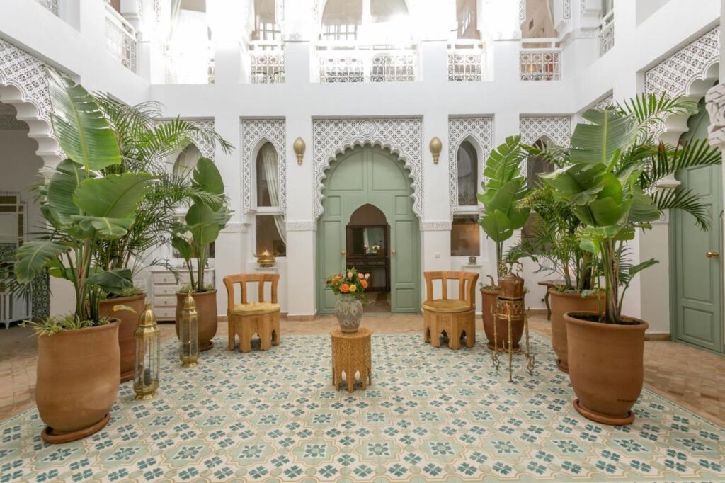 12 Charming Budget Riads in Marrakech 38 A bright, spacious room in one of the budget riads in Marrakech features intricate white arches, green doors, large potted plants, wooden chairs, and patterned tiles on the floor for an elegant and airy atmosphere.
