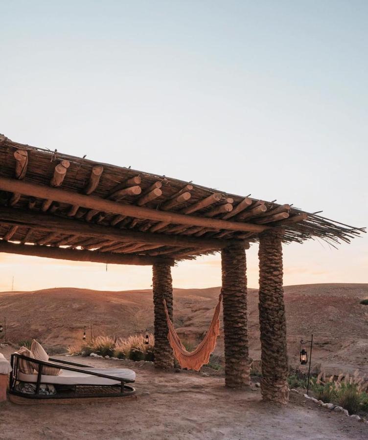 8 SPECTACULAR Agafay Desert Camps 11 An outdoor lounge with a rustic wooden pergola, hammock, and cushioned seating overlooks the Agafay desert camps at sunset, offering stunning views of the landscape beneath a clear sky.