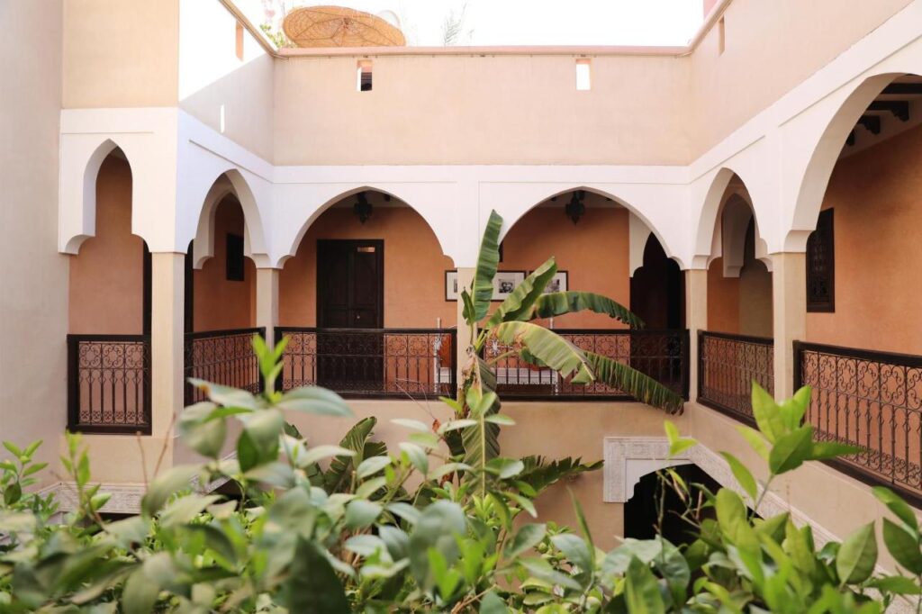 12 Charming Budget Riads in Marrakech 23 A view of a courtyard in traditional Moroccan style, surrounded by arched walkways and beige walls—typical of many charming budget riads in Marrakech—features leafy green plants and decorative railings in the tranquil foreground.