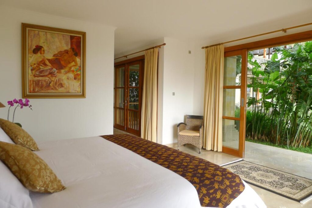 13 Most UNIQUE Places to Stay in Ubud 50 A bright bedroom with a made bed, gold-patterned pillows and runner, large painting above the headboard, sliding glass doors with cream curtains, a wicker chair, and views of lush greenery—one of the unique places to stay in Ubud.