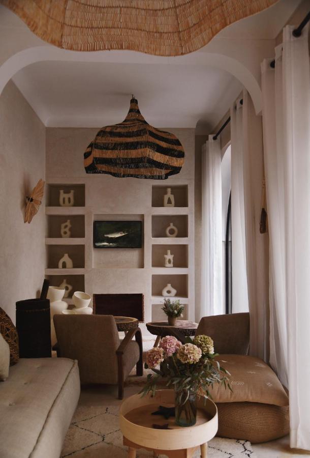 12 Charming Budget Riads in Marrakech 37 A cozy living room inspired by budget riads in Marrakech, with neutral tones, woven decor, modern furniture, a unique striped pendant light, open shelves with decorative items, large windows with sheer curtains, and a bouquet of flowers on a round wooden table.