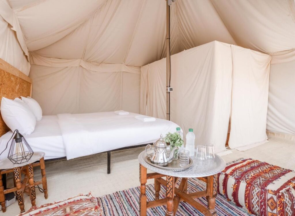 8 SPECTACULAR Agafay Desert Camps 38 Interior of a glamping tent at Agafay Desert Camps with a neatly made bed, two small wooden side tables, a tray with a teapot, glasses, and a water bottle on a round table, and patterned rugs adding charm to the floor.