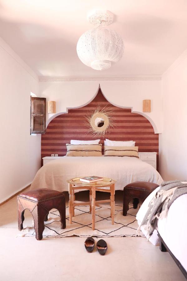 12 Charming Budget Riads in Marrakech 22 A cozy bedroom inspired by budget riads in Marrakech features a large bed, decorative wooden headboard, round mirror, two small stools, a wooden table, patterned rug, and a white pendant light. Slippers rest on the floor near the bed’s edge.
