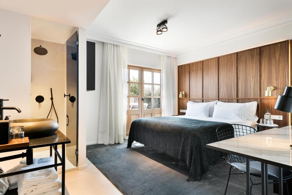 Top Barcelona Hotels with Rooftop Pool 10 Modern hotel room with a large bed, dark bedding, wooden headboard, and desk. An open bathroom with black sink adds style, while sunlight streams in. Perfect for those seeking Barcelona hotels with rooftop pool for relaxation.