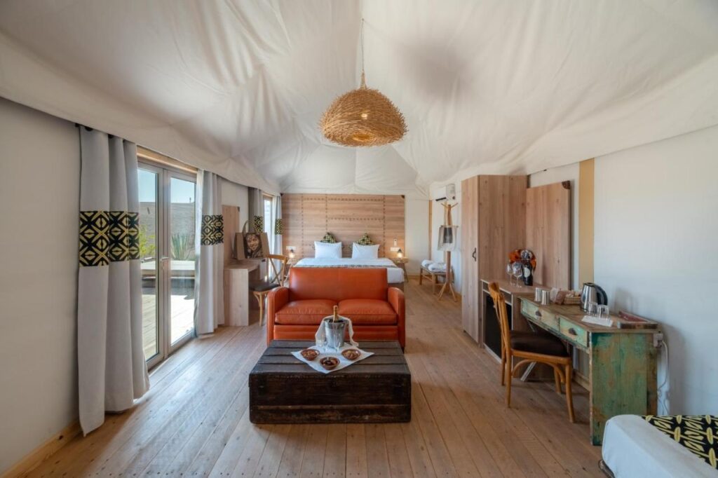8 SPECTACULAR Agafay Desert Camps 31 A spacious, bright glamping tent interior inspired by Agafay Desert camps, with a double bed, leather sofa, rustic wooden chest as a coffee table, desk, large windows for natural light, earthy decor, and a woven pendant light.