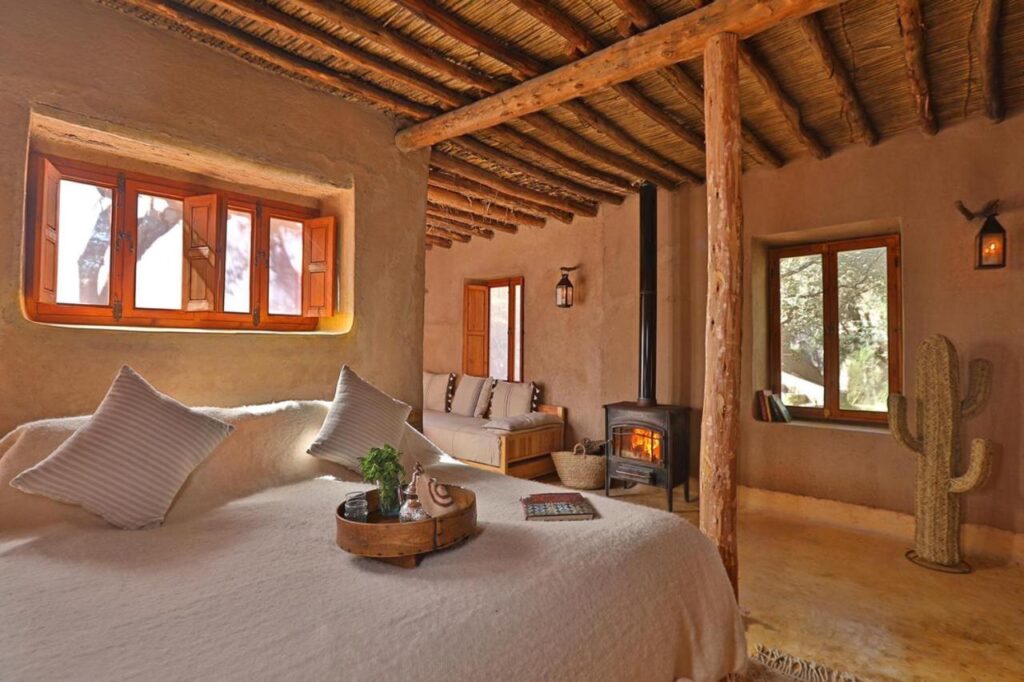 8 SPECTACULAR Agafay Desert Camps 12 Cozy rustic bedroom with wooden beams, a large bed with white linens, a wooden tray with plants, a corner wood stove, and a window seat. The warmth and inviting style echoes the tranquil charm of Agafay Desert Camps.