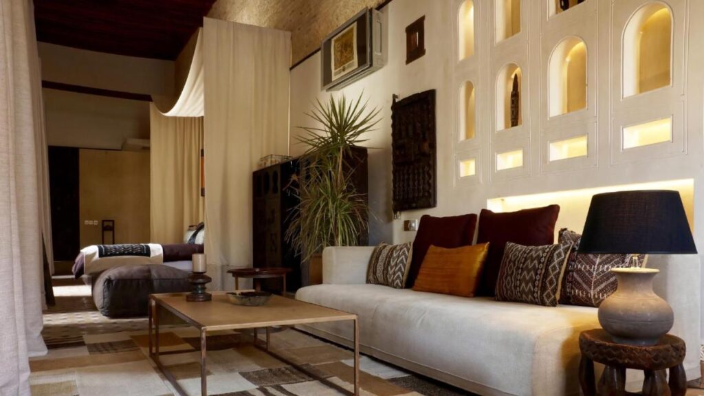 7 UNIQUE Riads in Fez You Won't Forget 7 A stylish living space inspired by riads in Fez, with a cream sofa, patterned cushions, black lamp, coffee table, indoor plant, and softly lit arched niches. A bedroom with twin beds is visible in the background, separated by a curtain.