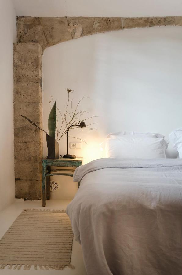 Unique Agroturismos in Mallorca 14 A cozy bedroom with white walls and bedding, a rustic stone arch reminiscent of agroturismos in Mallorca, a weathered wooden bedside table with a lamp, wildflowers in a vase, and a small woven rug on the floor.