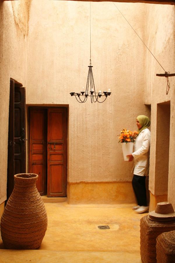 8 SPECTACULAR Agafay Desert Camps 14 A person in a light green headscarf and white coat carries a white pot of orange flowers through a sunlit, tan-walled room inspired by Agafay Desert Camps, with a woven basket, wooden door, and black chandelier.