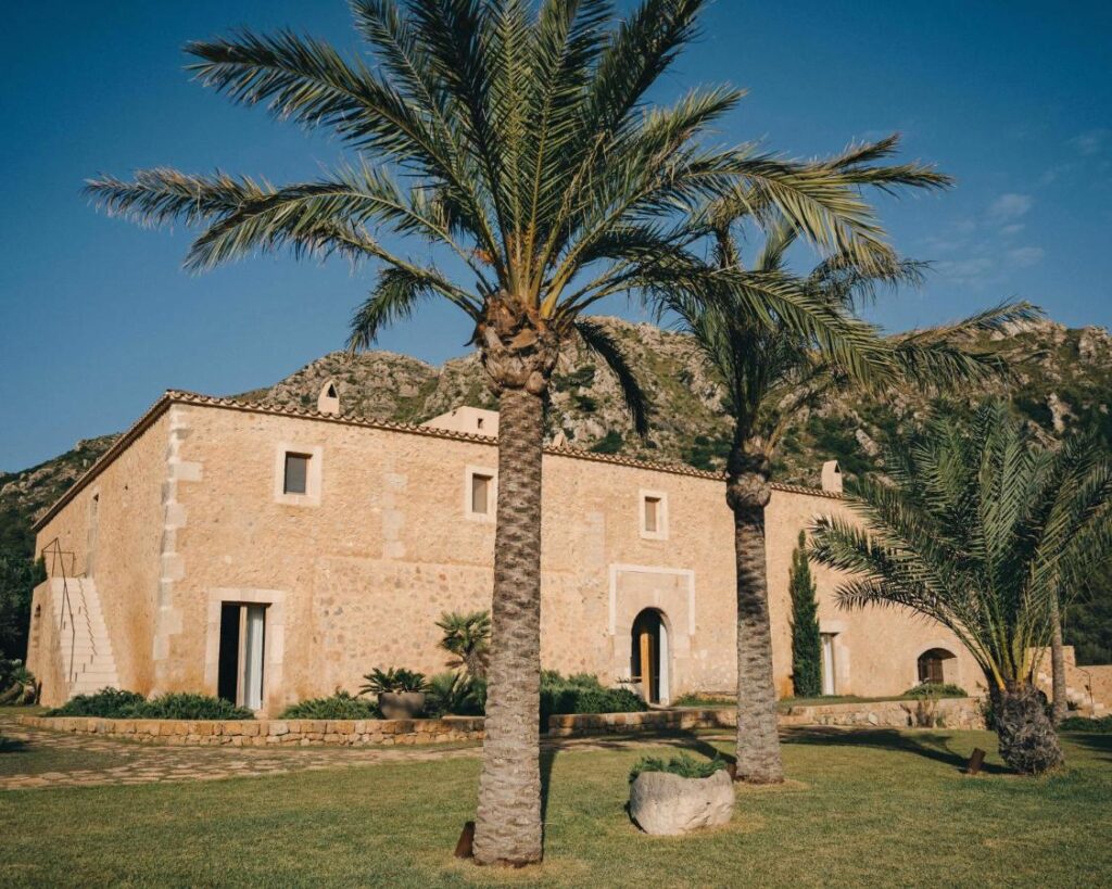 Unique Agroturismos in Mallorca 25 A large stone building with rectangular windows and an arched doorway stands among palm trees, set against a backdrop of mountains and a clear blue sky—an inviting example of agroturismos in Mallorca.