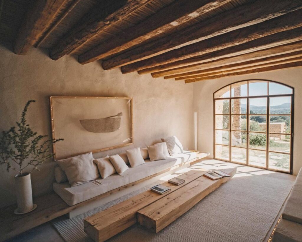Unique Agroturismos in Mallorca 22 A sunlit rustic living room, inspired by agroturismos in Mallorca, features a long white sofa with cushions, a low wooden coffee table, a large arched window, exposed wooden beams, and minimalist decor.