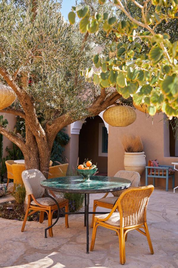 8 SPECTACULAR Agafay Desert Camps 23 A cozy outdoor patio with a round green table, three wicker chairs, and a fruit basket, set under a leafy tree. Warm sunlight filters through woven lanterns above, evoking the relaxed elegance of agafay desert camps.