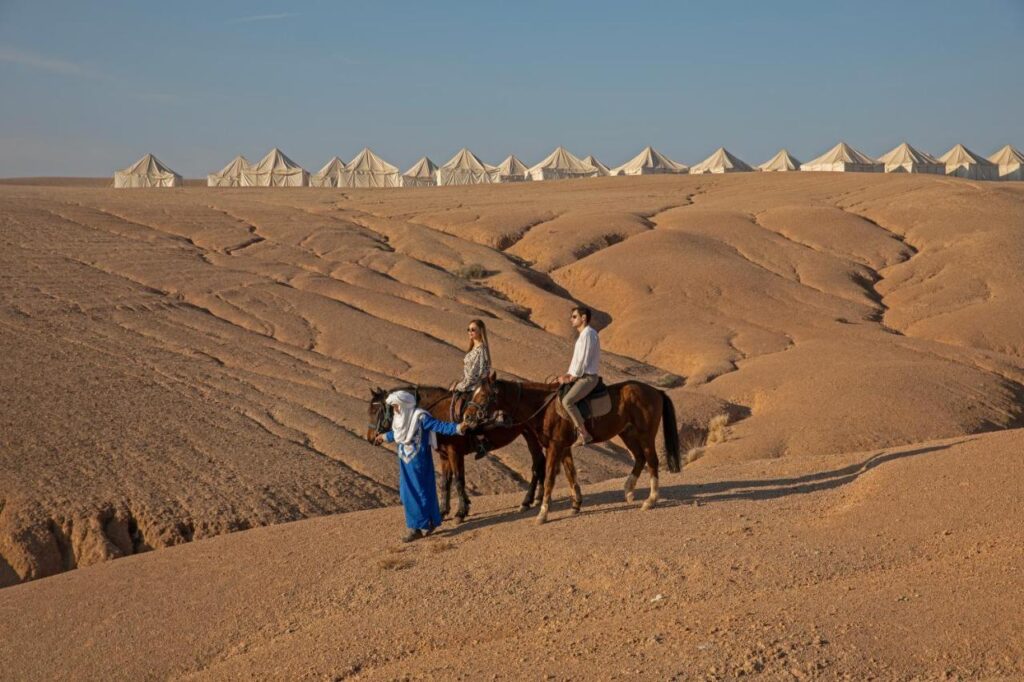 8 SPECTACULAR Agafay Desert Camps 36 Three people, two riding horses and one leading on foot, journey across a barren, sandy expanse near agafay desert camps. In the background, rows of white tents stand out against the clear blue sky.