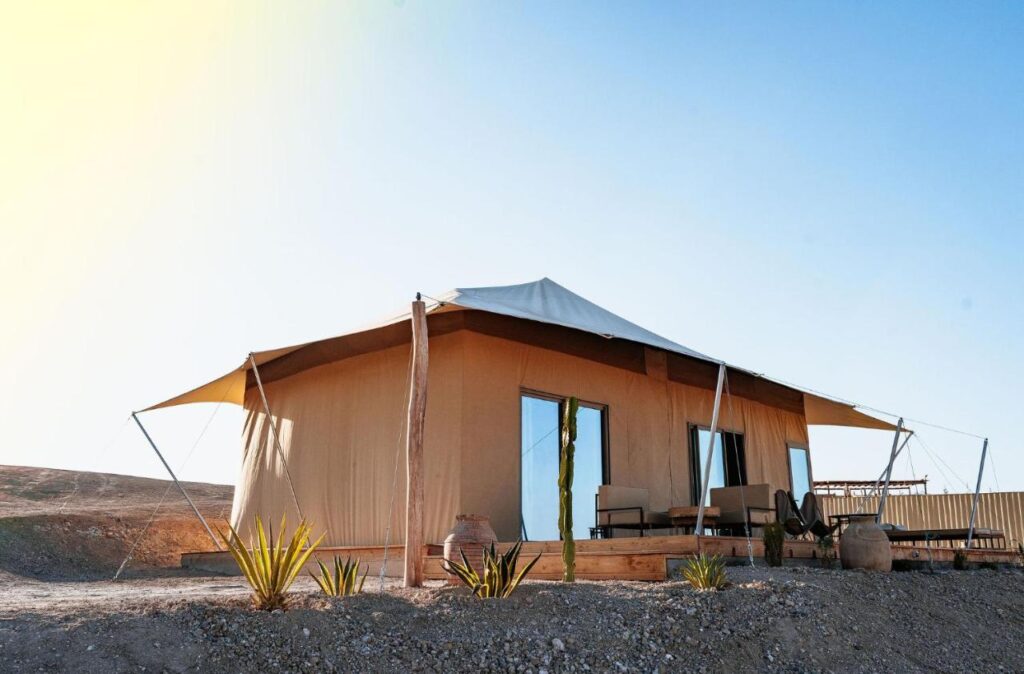 8 SPECTACULAR Agafay Desert Camps 33 A modern canvas tent with large windows and an outdoor seating area stands on a wooden deck in a desert landscape under a clear blue sky, capturing the essence of Agafay Desert camps. Small plants and pottery decorate the sandy foreground.