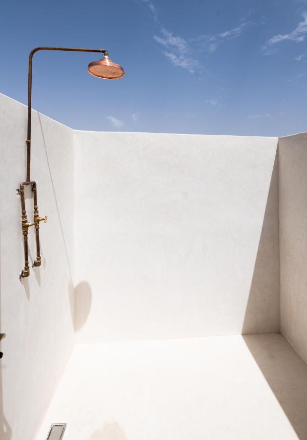 8 SPECTACULAR Agafay Desert Camps 34 Outdoor minimalist shower with a copper showerhead and exposed piping on white walls, reminiscent of agafay desert camps. Under a clear blue sky, sunlight casts sharp shadows across the clean, simple space.