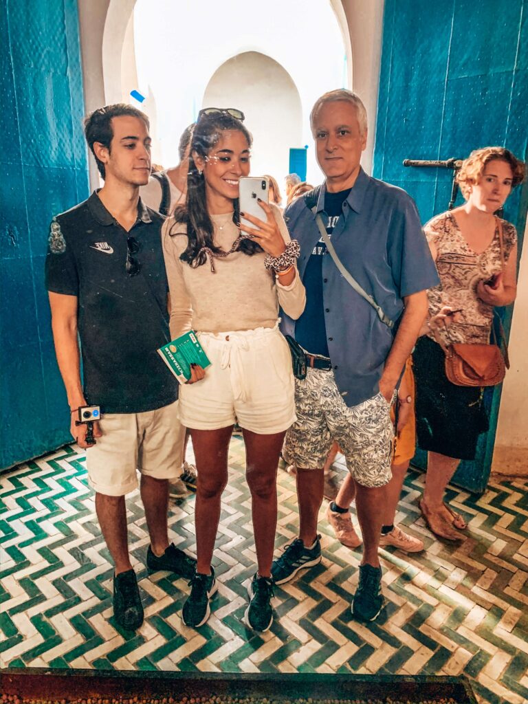 10 Unique Riads in Marrakech with Pool 45 Three people stand together taking a mirror selfie in a bright room with blue walls and patterned tile floors, typical of budget riads in Marrakech. A woman in the center holds the phone, with a man on either side. Other people are visible in the background.