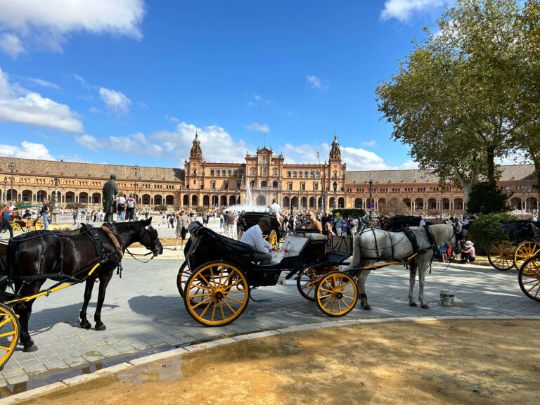 Horse-drawn carriages with yellow wheels wait in front of the grand, ornate Plaza de España in Seville, Spain—an iconic stop for anyone spending 1 day in Seville, Spain—as people stroll around the large plaza on a sunny day.