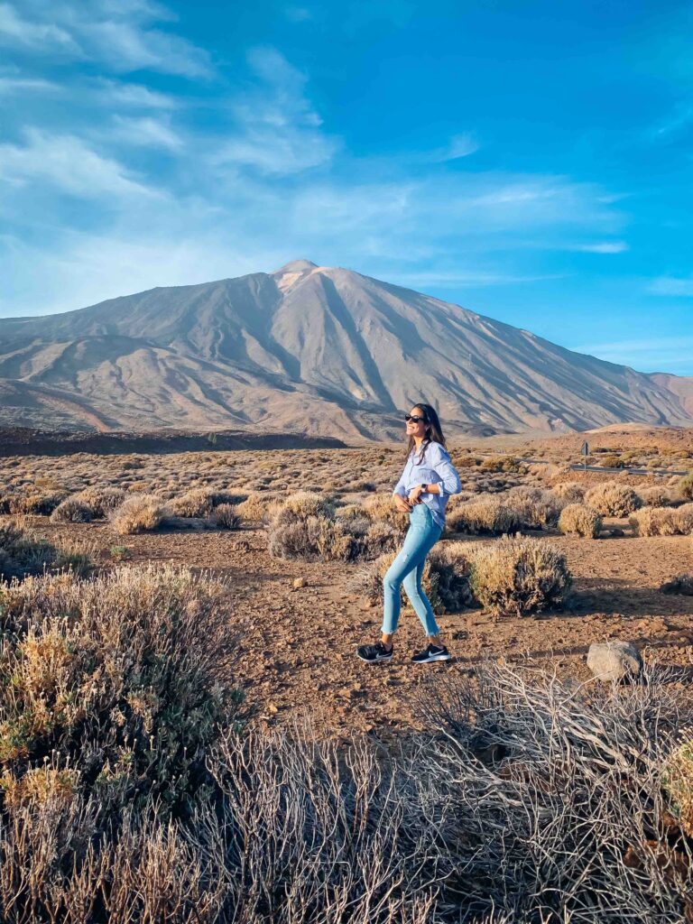 11 UNIQUE Boutique Hotels in Tenerife 4 A woman wearing sunglasses, a white shirt, and light blue jeans stands in a dry, rocky landscape with shrubs, framed by a large mountain and blue sky—capturing the adventurous spirit of those seeking boutique hotels in Tenerife.